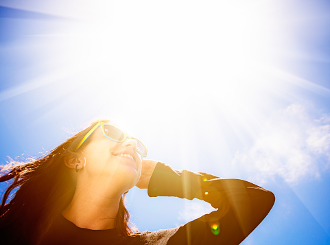 A pretty young brunette in sunglasses stands outdoors looking up, her hand raised to shield her eyes from the dazzling sun. Lots of copy space over the sun.