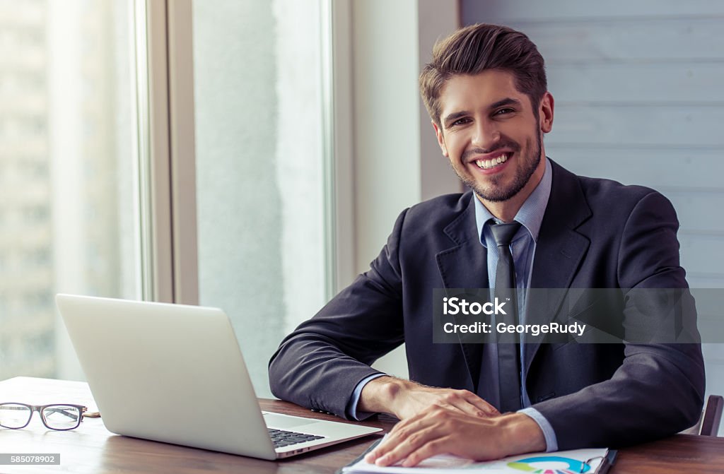 Handsome young businessman working Portrait of handsome young businessman in formal suit using a laptop, looking at camera and smiling while working in office Insurance Agent Stock Photo