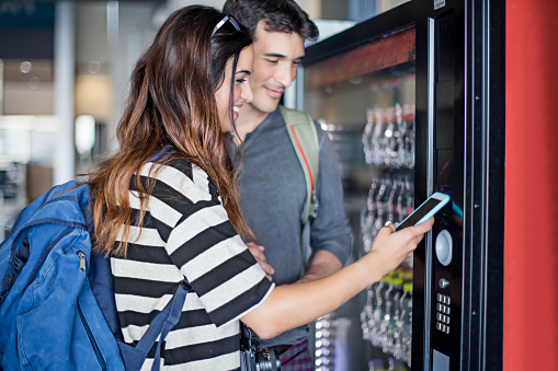 Young couple paying with mobile phone the soft drink at vending machine in the airport.
