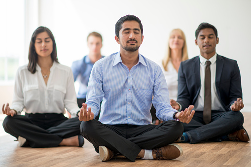 A multi-ethnic group of corporate business professionals are meditating together while wearing business attire. They are sitting with their eyes closed and are silently meditating.