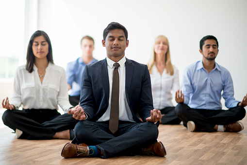 A multi-ethnic group of corporate business professionals are meditating together while wearing business attire. They are sitting with their eyes closed and are silently meditating.