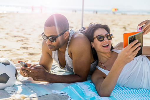 A photo of couple wearing sunglasses relaxing at beach. Man is using phone and woman reading book. They are wearing casuals.
