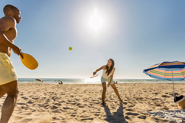 Happy couple playing tennis at beach on sunny day A photo of happy couple playing tennis. Woman enjoying with man at beach. They are against clear sky. racquet stock pictures, royalty-free photos & images