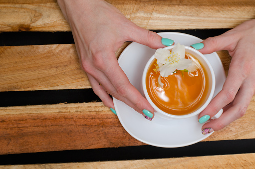 Women's hands with mug of green tea and jasmine flower on the background of wooden planks, top view