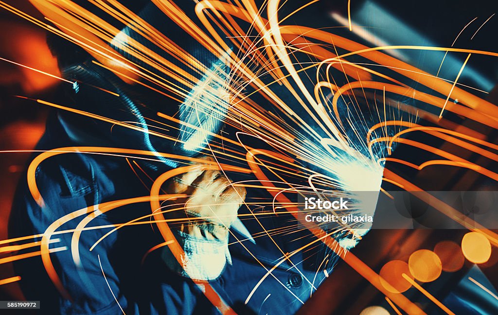 Welding two pieces of metal. Unrecognizable man welding metal plate and construction frame. This is arc welding.The worker is using protective mask and blue working uniform. Background is lit orange. Long exposure. Sparks Stock Photo