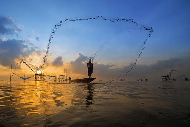 Asian fisherman on wooden boat casting a net for catching freshwater fish in nature river in the early morning before sunrise at Pak Pra, Phatthalung, Thailand.