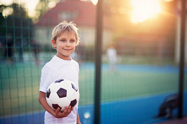 Little boy playing football in the schoolyard Little boy playing football in the schoolyard. The boy aged 7 is holding the football and smiling. Schoolyard and the setting sun in the background. recess soccer stock pictures, royalty-free photos & images