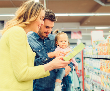 Young family with one child in groceries shopping. Father carry his baby daughter. They are in supermarket buying groceries. Choosing baby food. Woman holding box and they are reading nutrition label.