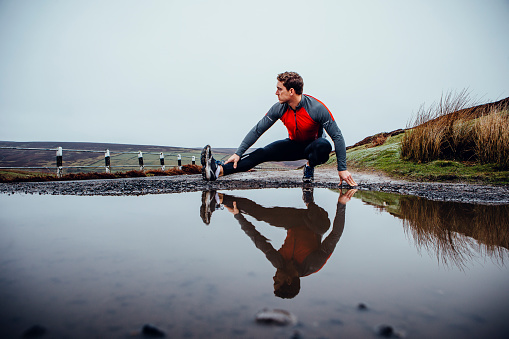 Male runner training outdoors in wet weather. His reflection seen in a puddle of water before him. He stretched his leg.