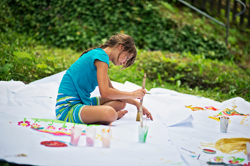 Little girl having fun painting. Giant sheets of paper are laid down on grass, the girl is sitting on the paper and painting. Girl is aged 10.