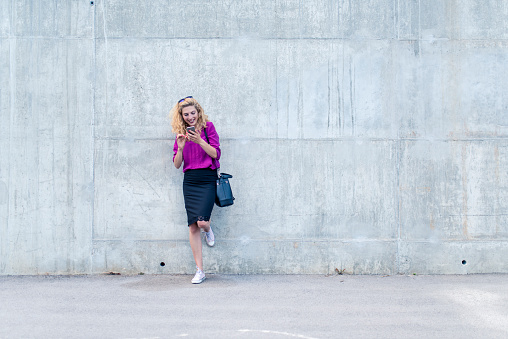 Businesswoman standing in  front of concrete wall, using her mobile phone. She is happy and she is smiling.