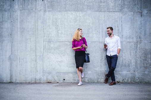 Businessman and businesswoman standing in  front concrete wall of office building and using mobile phones. They are smiling and looking at each other.