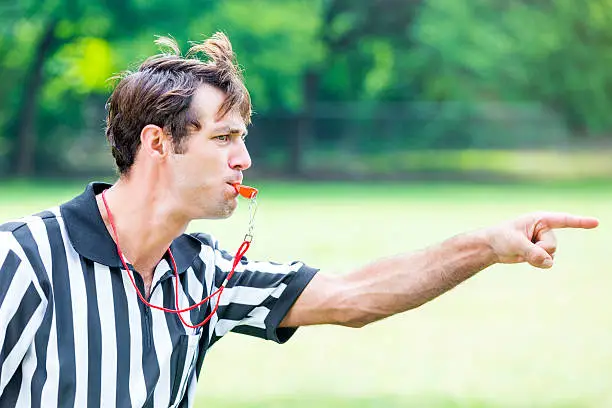 Handsome mid adult Caucasian referee points as he blows his whistle. He is calling a penalty or foul during sporting event. He has brown hair and is wearing a black and white striped referee uniform. He is standing on the playing field. He has an intense expression on his face.