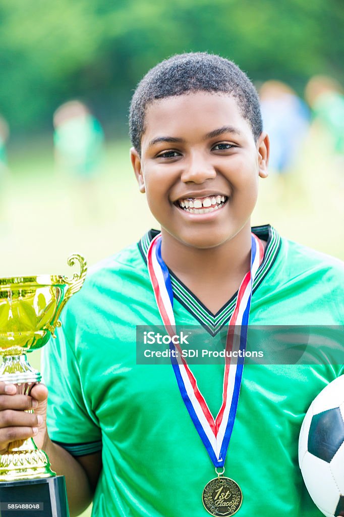 Excited African American teenage soccer player with championship trophy Handsome teenage African American soccer player holds his team's championship golden trophy cup. He is also wearing a medal around his necck and holding a soccer ball. He is smiling proudly. He is wearing a green jersey. He has short black hair. His teammates are blurred in the background. Active Lifestyle Stock Photo