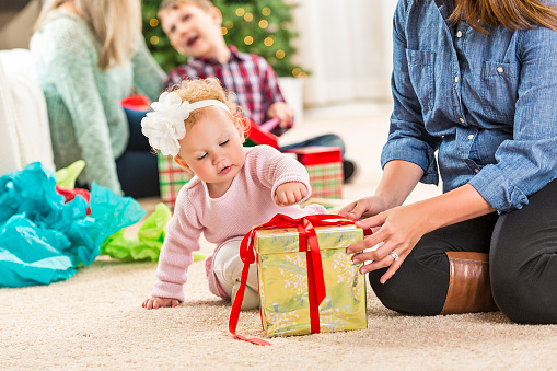 Adorable baby girl curiously reaches for Christmas gift. Her mother is sitting on the floor with her holding the gift. The baby is wearing pink pajamas and has curly blond hair with a headband around her head. The mom is wearing a denim shirt, black pants and brown boots. The baby's brother and grandmother are opening gifts in the background.