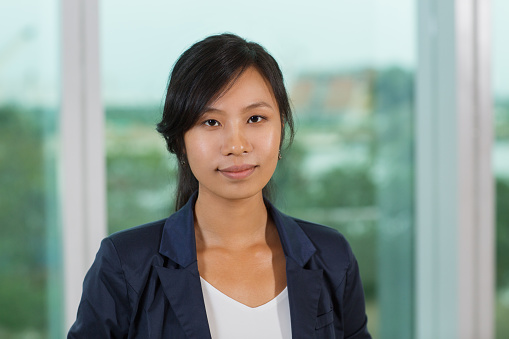 Close up portrait of slightly smiling young businesswoman and looking straight with window and blurry city view outside in background