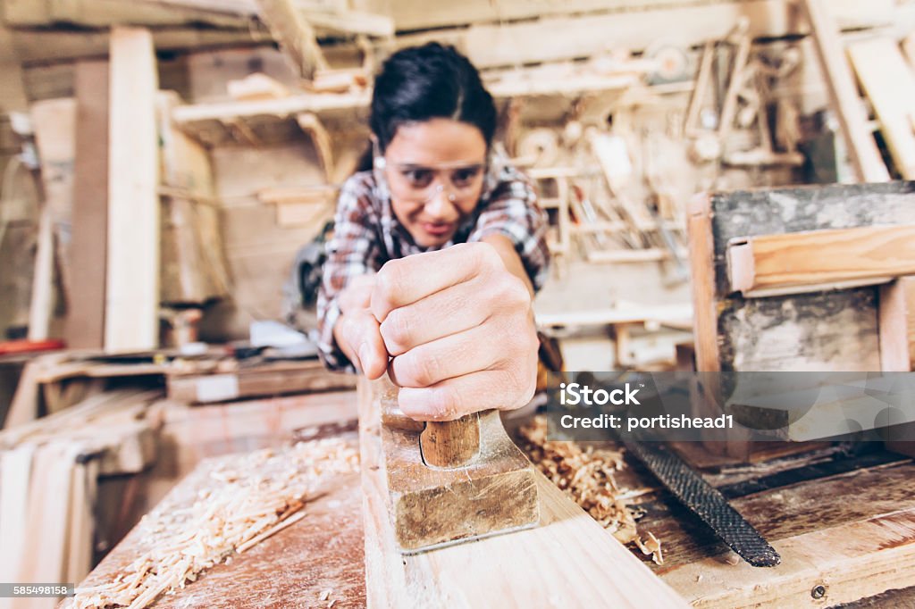 Concentrated female carpenter using grater at workshop Concentrated young female carpentry manual worker using grater at her workshop. Tools on shelves and wooden material on background. Focus on foreground. One Woman Only Stock Photo