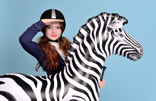 redhead girl with freckles in riding clothes and helmet on a light background with a bouncy horse, the jockey concept, concept dream