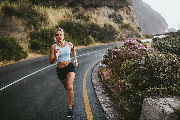 Fitness woman running on countryside highway Full length shot of young fitness woman running outdoors on an open highway in countryside. Determined female athlete sprinting on road during rain. hard and fast stock pictures, royalty-free photos & images