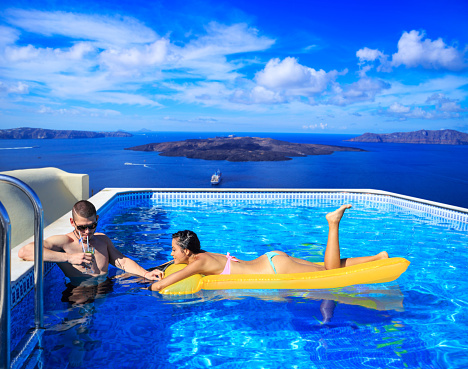 Young couple having fun at swimming pool. Man with sunglasses drinking coctail, woman leaning on yellow inflatable beach mattress. Panoramic view of Santorini volcano island and yachts on background.
