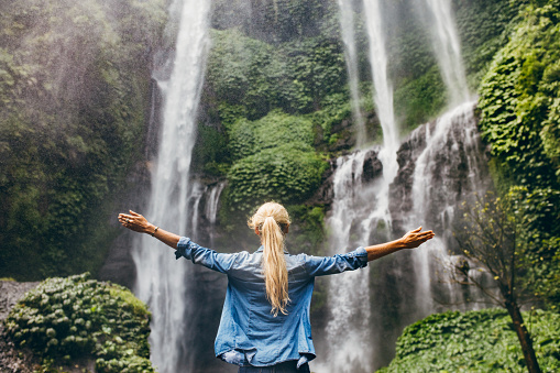 Rear view of young woman standing in front of waterfall with her hands raised. Female tourist with her arms outstretched looking at waterfall.