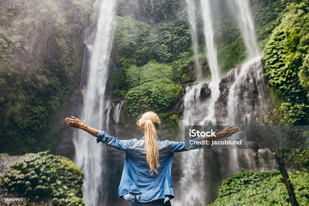 Woman standing by waterfall with her hands raised Rear view of young woman standing in front of waterfall with her hands raised. Female tourist with her arms outstretched looking at waterfall. Waterfall Stock Photo