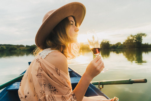 Young Caucasian woman in hat sitting in the boat and drinking wine 