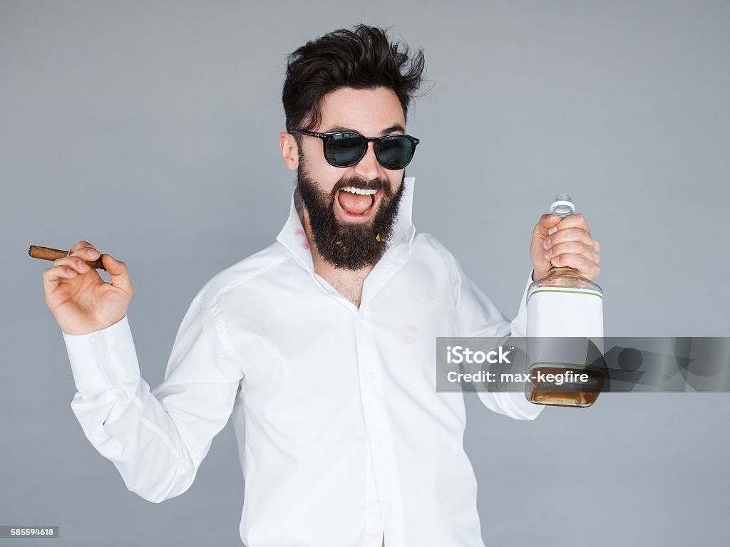 man holding a bottle of whiskey and cigar Work hard play hard. Handsome young man in sunglasses and white shirt holding a bottle of whiskey and cigar and shouting while standing against grey background Men Stock Photo