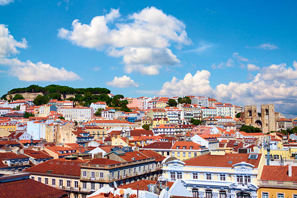 View across the city of Lisbon, Portugal on a summer day. The castle of Saint George is on the hill in the background, the historic downtown in the foreground and a lovely blue sky with fluffy clouds overhead. The cathedral is on the right side of the picture.