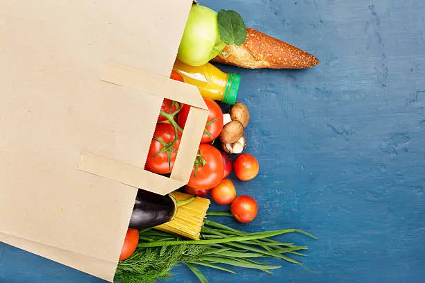 Paper bag with different of vegetables and fruits on blue surface with copy space, top view