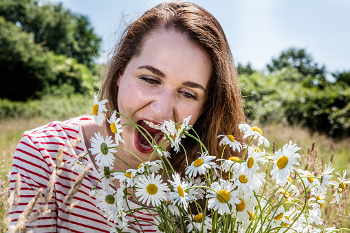 beautiful young woman enjoying eating camomile field flowers for fun, smiling for natural wellbeing in summertime daylight
