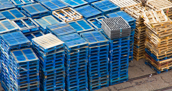 Horizontal image of wooden shipping pallets stacked on dock.  There are a few unpainted wooden shipping pallets as well as some grey plastic shipping pallets as well.