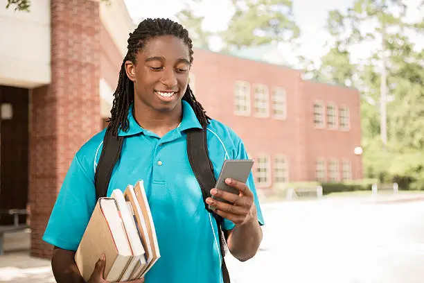 Back to school.   Handsome, African descent boy heads off to college.  The 18-year-old is excited to start his first day of school.  He carries a backpack and textbooks and texts using his cell phone.  College building in background. 