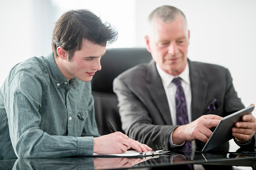 A student intern and businessman are working together in a corporate office to update the companies social media and check emails on a digital tablet.