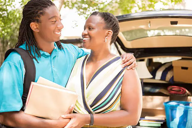 African descent boy heads off to college or moves away from home.  The 18-year-olds' mother is helping him pack up his car as he gets ready for the big move.  He is excited to start his college adventures and gives mom a big hug to say goodbye. He wears a backpack.  Family events.  Back to school.