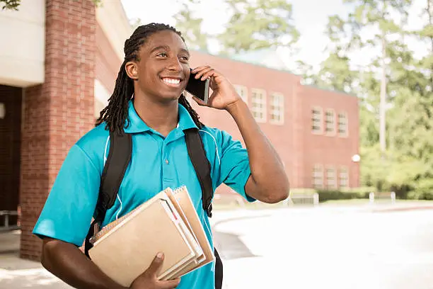 Back to school.   Handsome, African descent boy heads off to college.  The 18-year-old is excited to start his first day of school.  He carries a backpack and textbooks and talks on cell phone.  College building in background. 