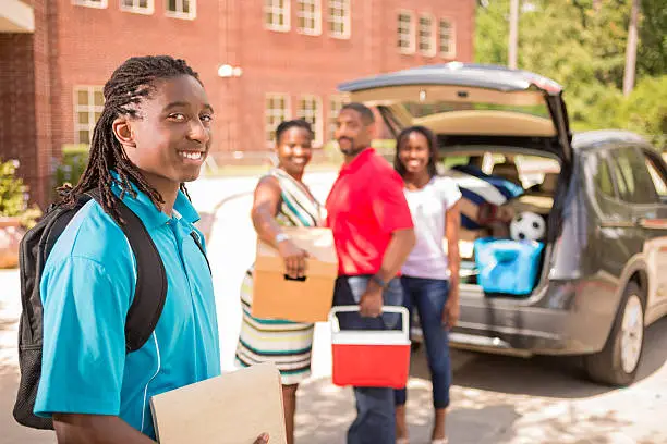 African descent boy heads off to college.  The 18-year-olds' family are all helping him unpack his car as he moves into the college campus dorm.  He is excited to start his school adventures. He  carries a backpack and textbooks.  Family events.  Back to school.