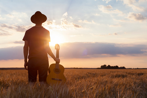 Musician holding acoustic guitar and walking in summer fields at sunset