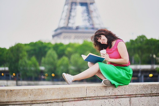 Cheerful young girl with coffee to go reading a book near the Eiffel tower. Student preparing for exam in Paris. International education exchange program in France concept