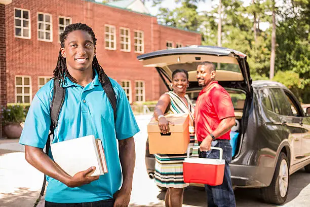 African descent boy heads off to college.  The 18-year-olds' parents are all helping him unpack his car as he moves into the college campus dorm.  He is excited to start his school adventures. He  carries a backpack and textbooks.  Family events.  Back to school.