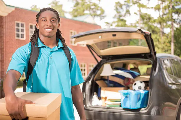 African descent boy heads off to college.  The 18-year-old is unpacking his car as he moves into the college campus dorm.  He is excited to start his school adventures. He carries a backpack and moving box.  Back to school.