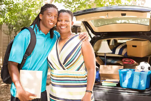 African descent boy heads off to college or moves away from home.  The 18-year-olds' mother is helping him pack up his car as he gets ready for the big move.  He is excited to start his college adventures and gives mom a big hug to say goodbye. He wears a backpack.  Family events.  Back to school.