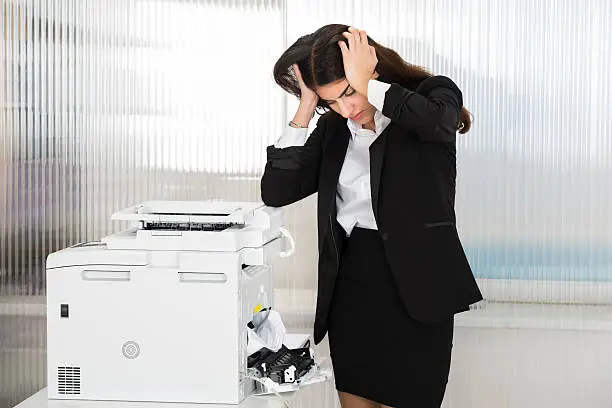 Irritated young businesswoman looking at paper stuck in printer at office