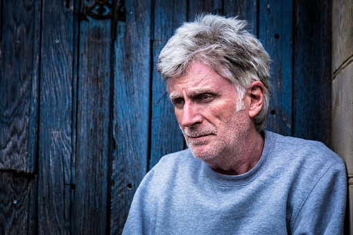 Medium close up image of a senior man with white/grey stubble sitting in a doorway with a sad, hopeless expression on his face. Horizontal colour image with copy space.