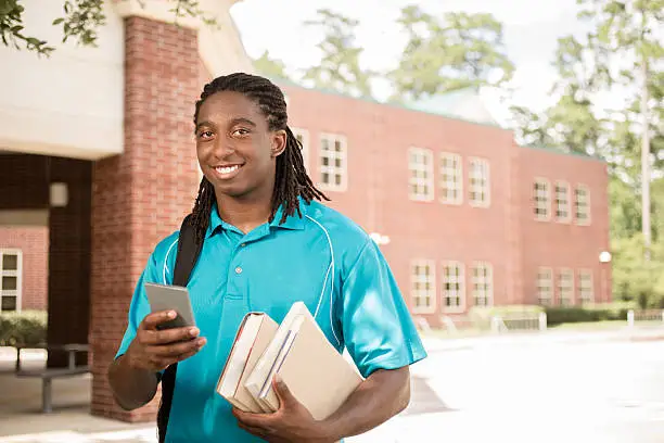 Back to school.   Handsome, African descent boy heads off to college.  The 18-year-old is excited to start his first day of school.  He carries a backpack and textbooks and texts using his cell phone.  College building in background. 