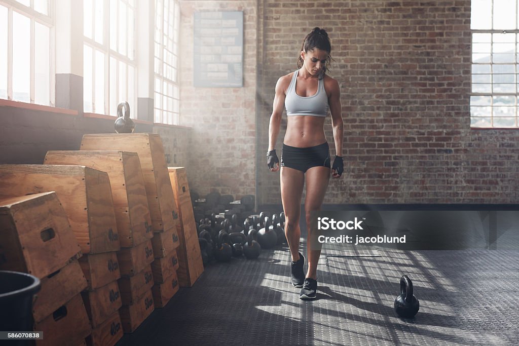 Fitness female getting ready for an intense workout Full length shot of young woman in sportswear taking a walk in the gym and looking at kettle bell on floor. Fitness female getting ready for intense workout. Kettlebell Stock Photo