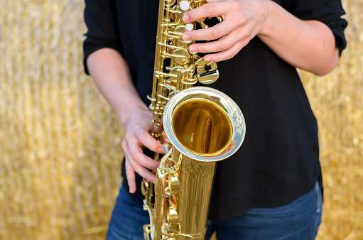 Close up on a shiny brass tenor saxophone being played by a young female musician standing in front of a hay bale outdoors