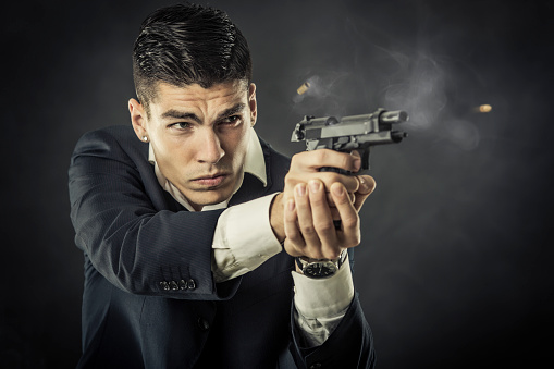 Young mafia man holding a pistol, shooting bullets. About 25 years old Caucasian male in suit.