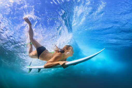 Young active girl in bikini in action - surfer with surf board dive underwater under breaking big ocean wave. Family lifestyle, people water sport adventure camp, beach extreme swim on summer vacation