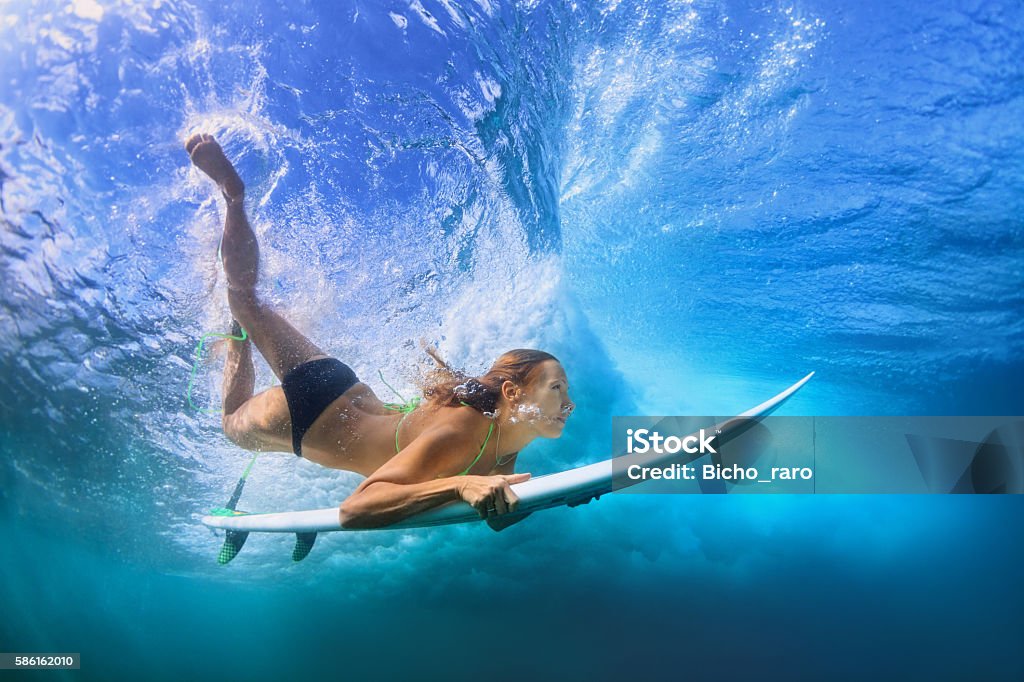 Beautiful surfer girl diving under water with surf board Young active girl in bikini in action - surfer with surf board dive underwater under breaking big ocean wave. Family lifestyle, people water sport adventure camp, beach extreme swim on summer vacation Surfing Stock Photo